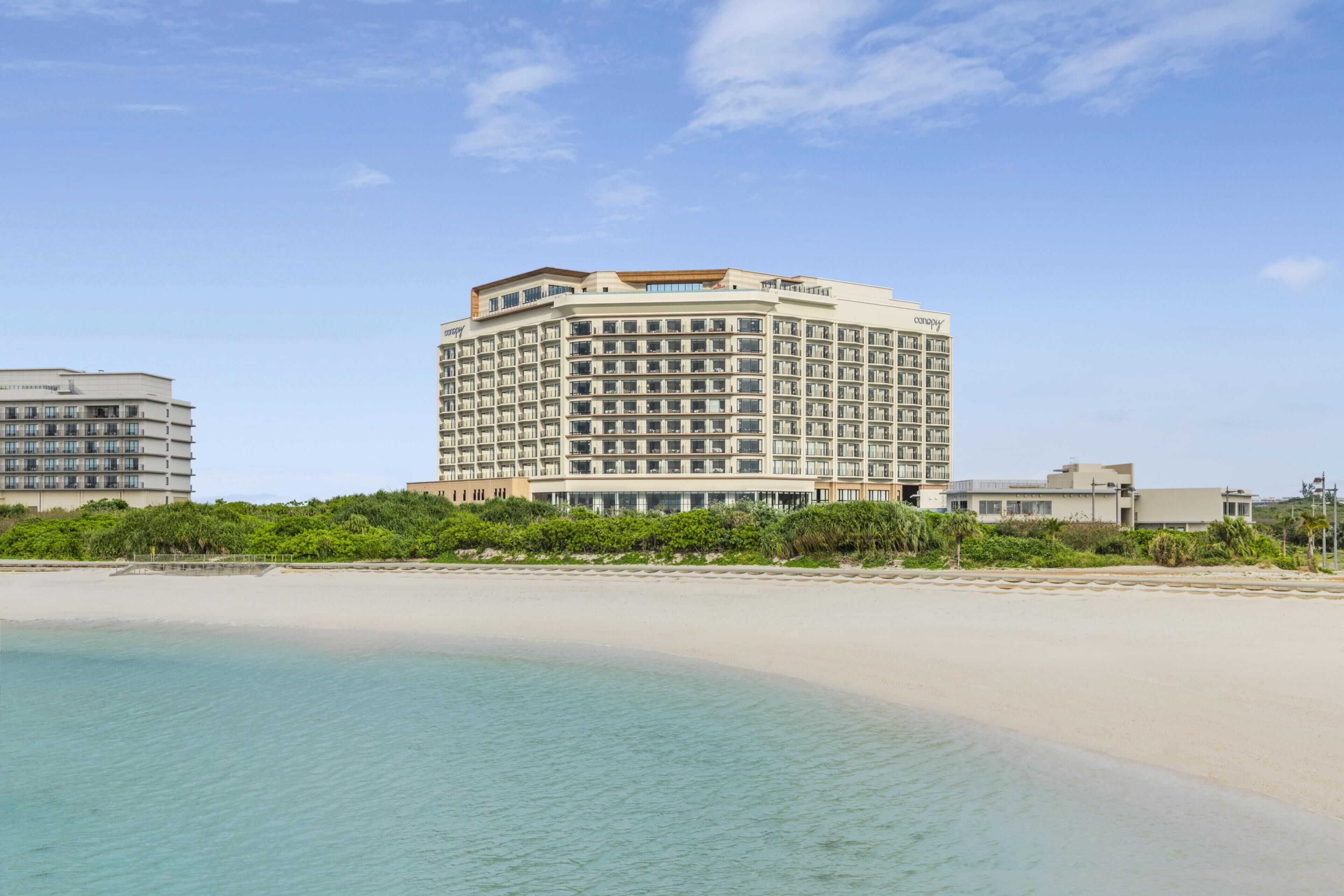 Un hotel frente a la playa con un edificio grande, una playa de arena y cielo azul despejado.