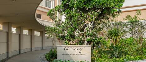 L’entrée d’un bâtiment, agrémentée d’une enseigne indiquant « Canopy Okinawa | Miyako Island Resort », est entourée de verdure et bordée d’un chemin sinueux.
