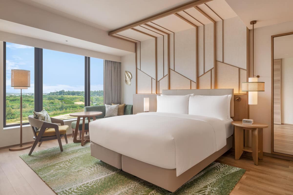 A modern hotel room with a large bed, a desk, a chair, and a view of the outdoors.