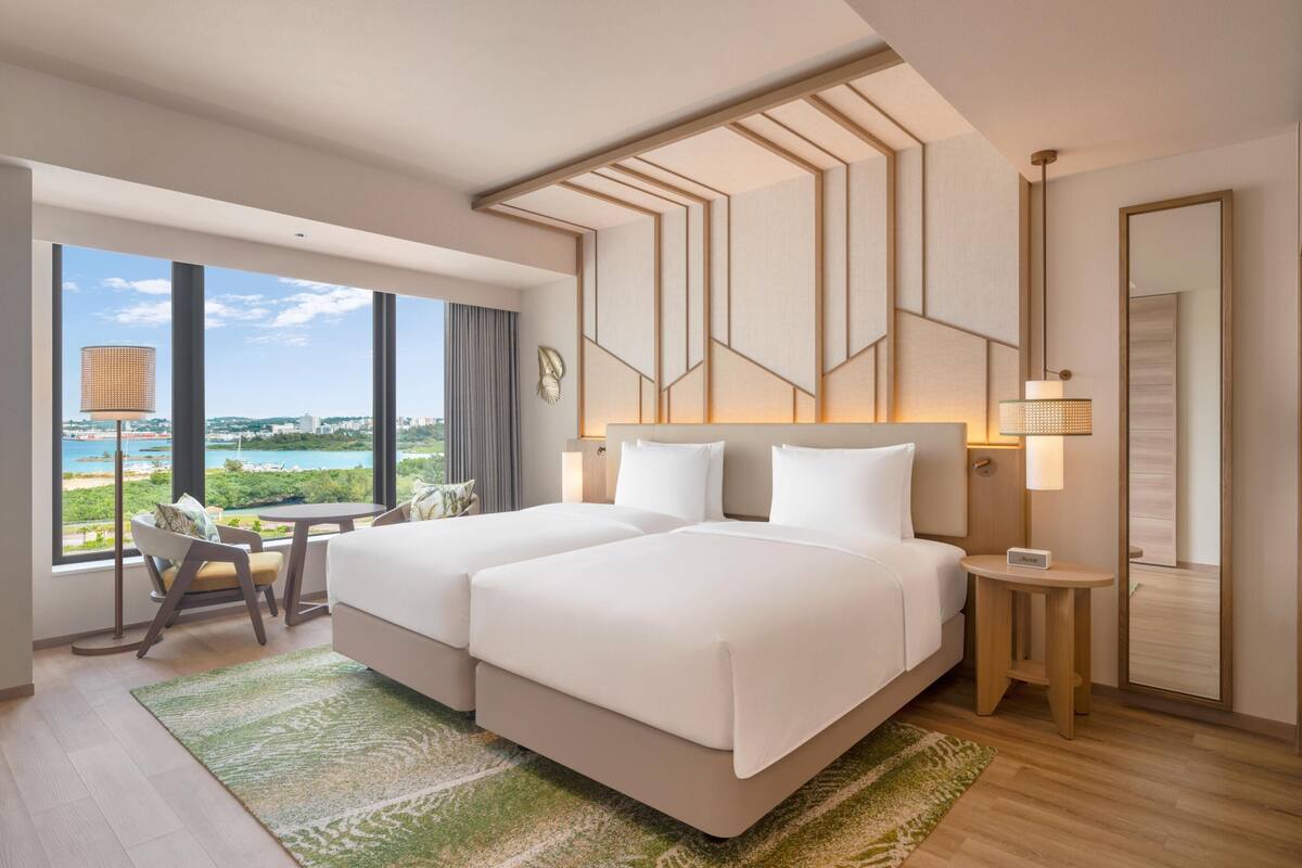 A hotel room with two beds, a view of the city, a small table, and a chair.