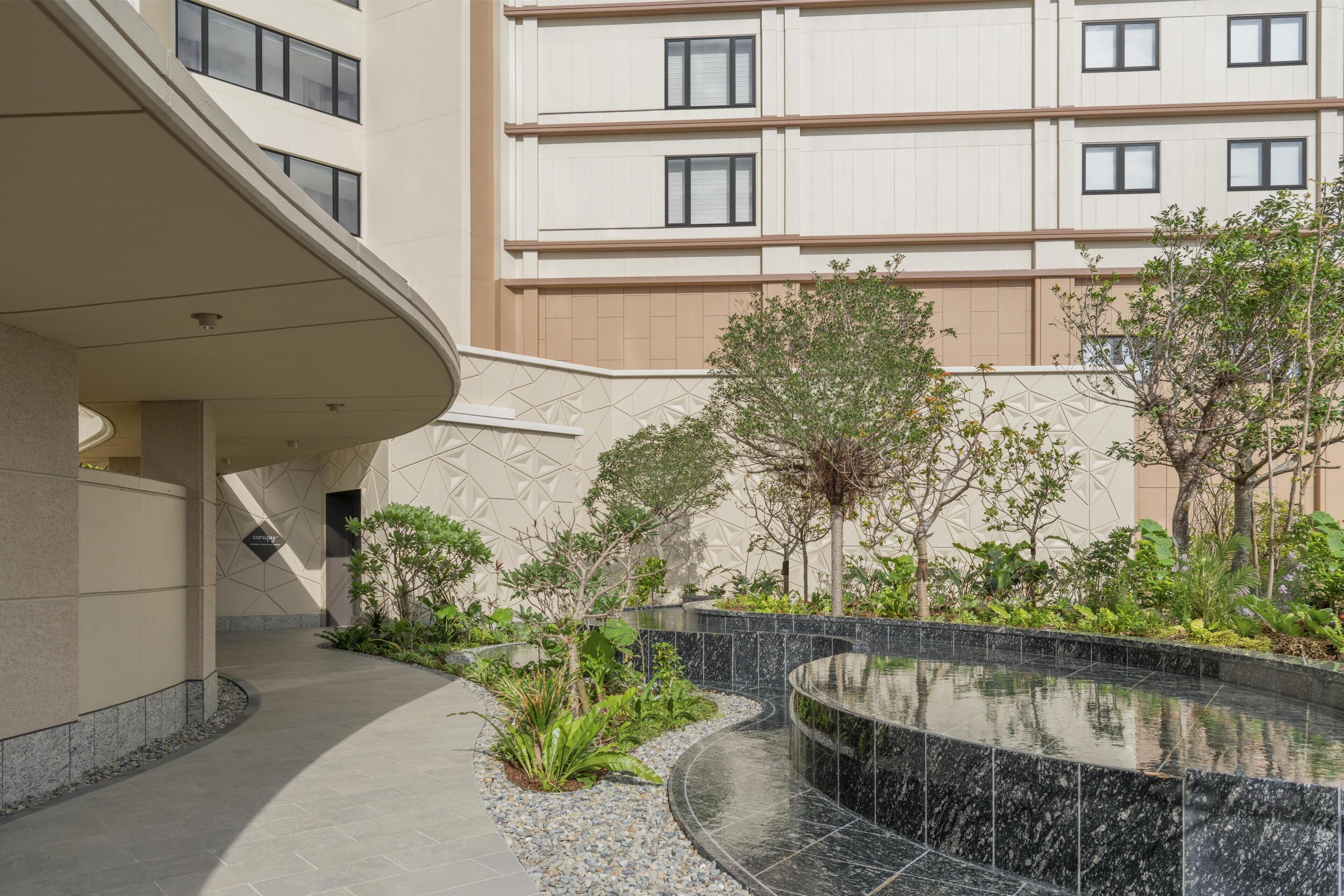 Un edifici modern amb una entrada corba, un jardí paisatjistic amb una font i una estructura de diversos pisos al fons.