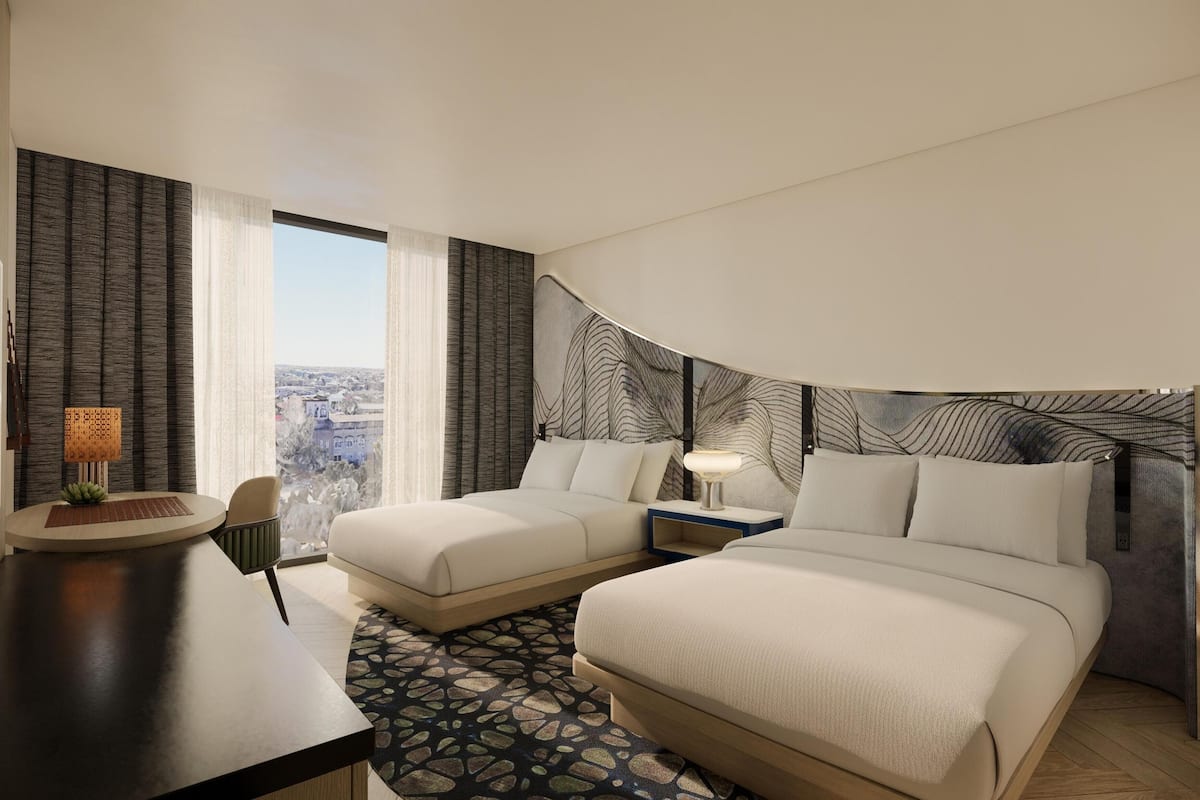 Habitación de hotel con dos camas, un escritorio, una silla, una lámpara y vista a la ciudad.