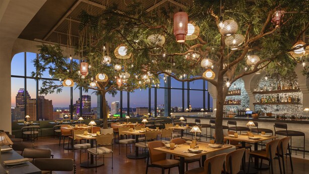 Un restaurante con mesas y sillas dispuestas bajo un árbol, con faroles colgantes, ventanas amplias con vista a la ciudad y una zona de bar con botellas y vasos.