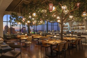 Un restaurant avec des tables et des chaises disposées sous un arbre, agrémenté de lanternes suspendues, de grandes fenêtres offrant une vue sur la ville et un bar avec des bouteilles et des verres.