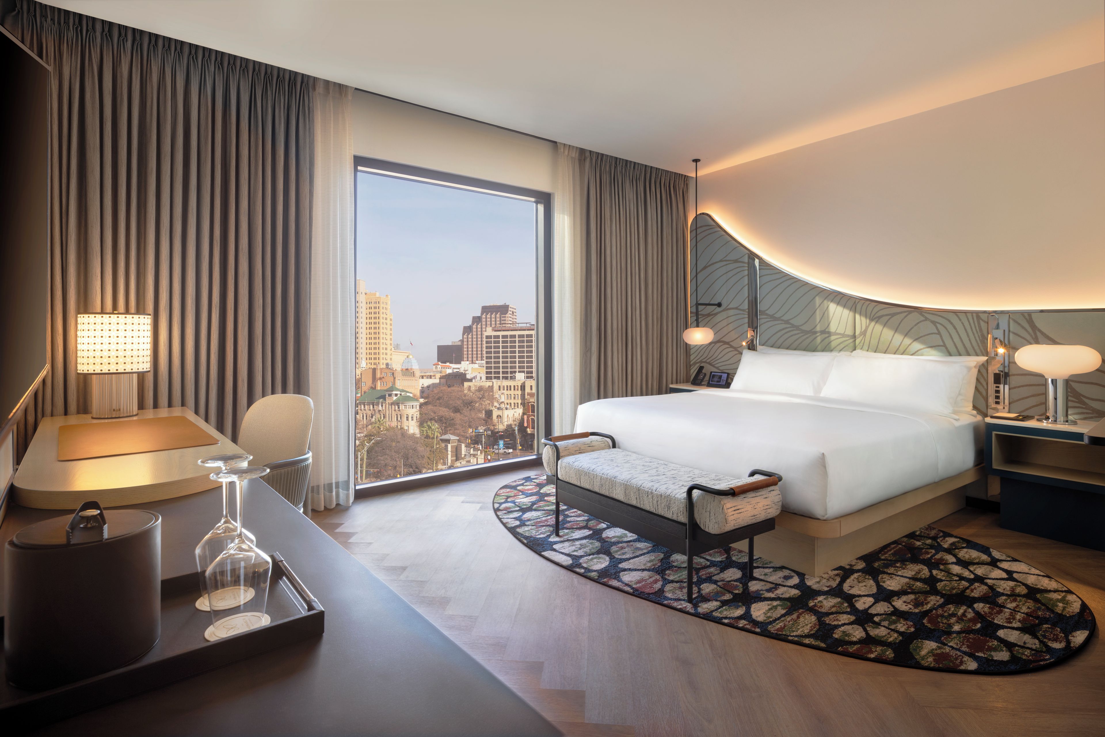A hotel room with a large bed, a desk with a lamp, a chair, a view of the cityscape through the window, and a patterned rug.
