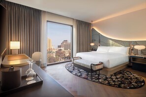 Een hotelkamer met een groot bed, een bureau met lamp, een stoel, en een uitzicht op de stad door het raam, plus een kamerbreed tapijt met dessin.