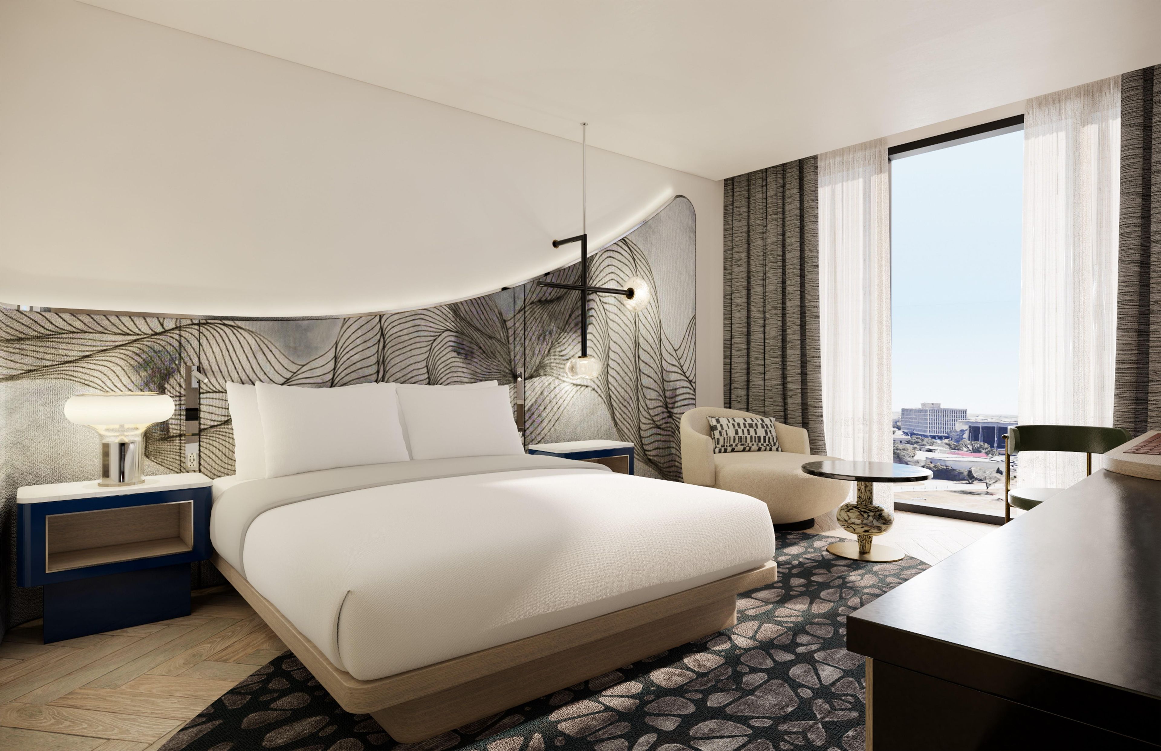 A modern hotel room with a large bed, a desk, a chair, a small table, and a view of the city through the window.