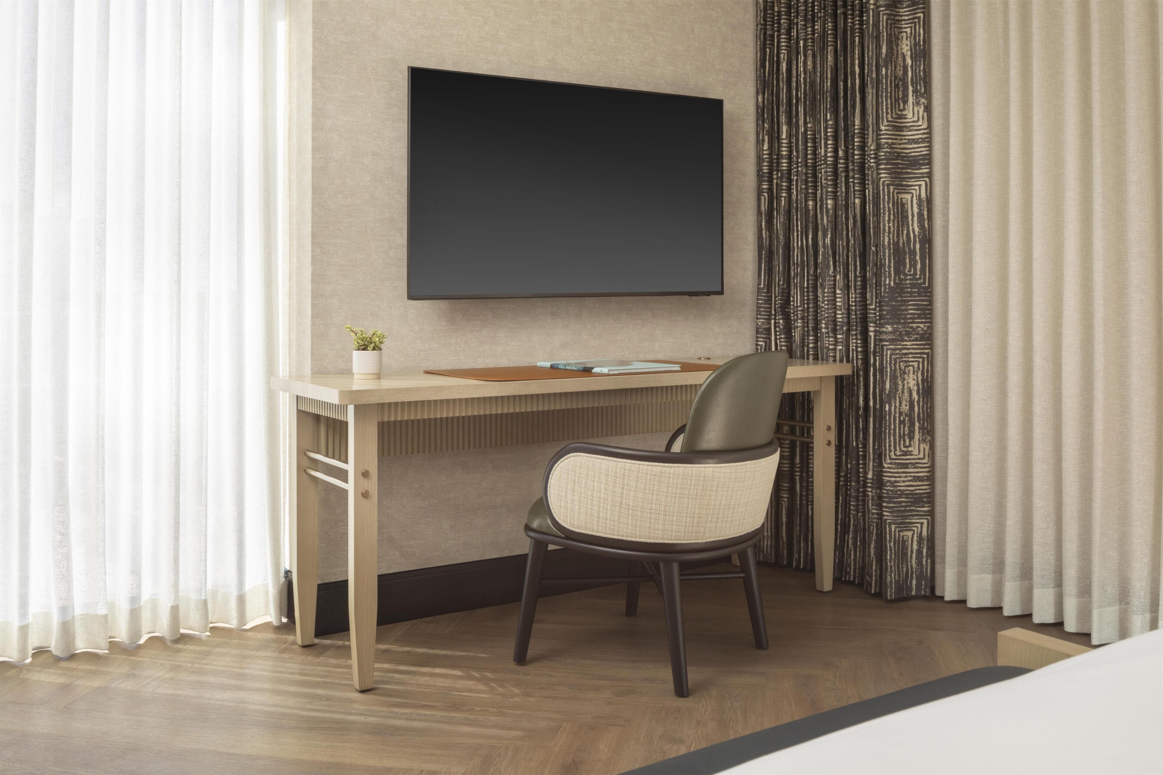 Ett hotellrum med ett skrivbord i trä, en stol, en platt-TV som är monterad på väggen och en krukväxt på skrivbordet.
