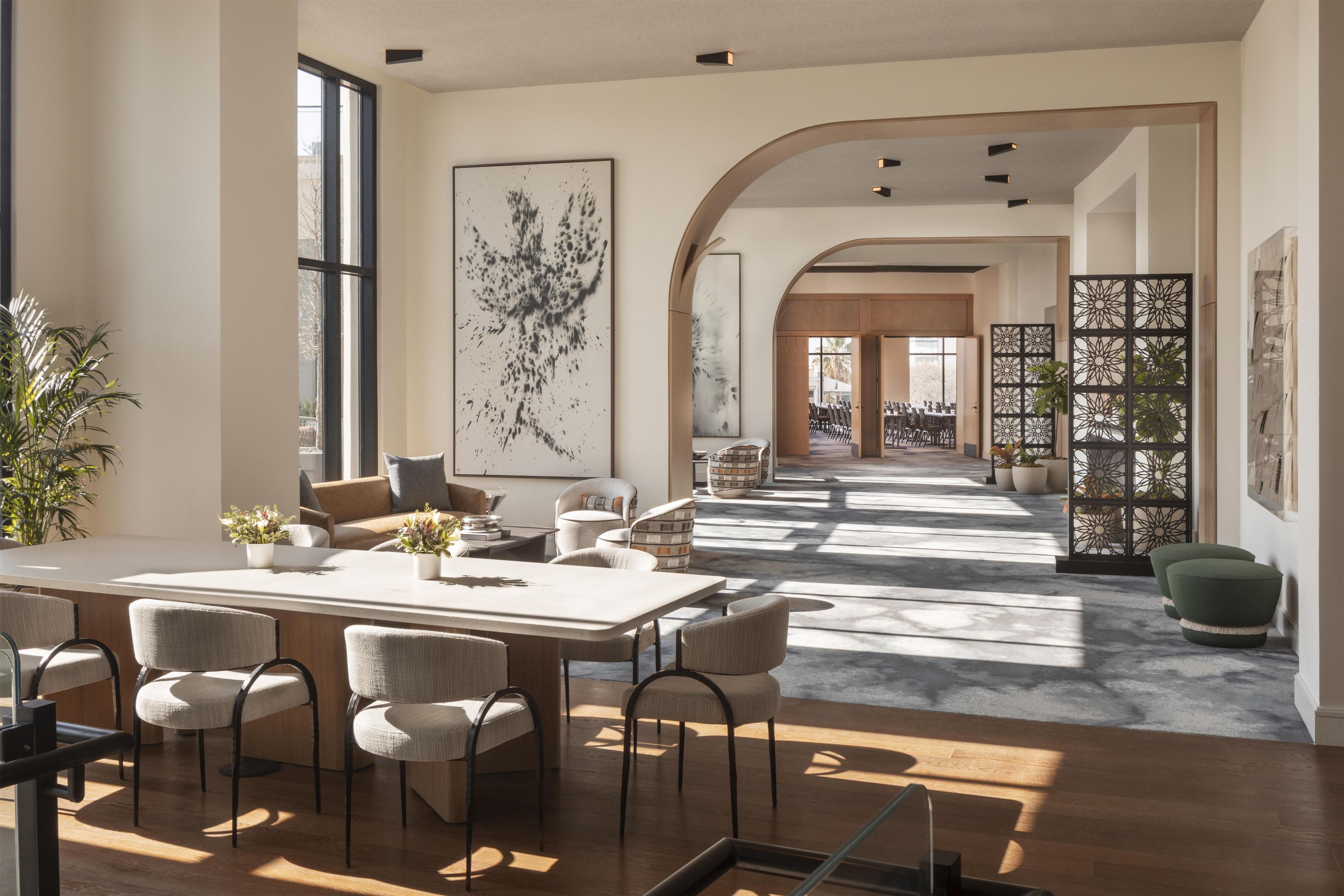 A spacious dining area with a long table and chairs, a large abstract painting on the wall, and a view of the dining area through an arched doorway.