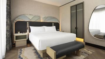 Una habitación de hotel con una cama, mesitas de noche, un banco, un espejo y un ventanal grande.
