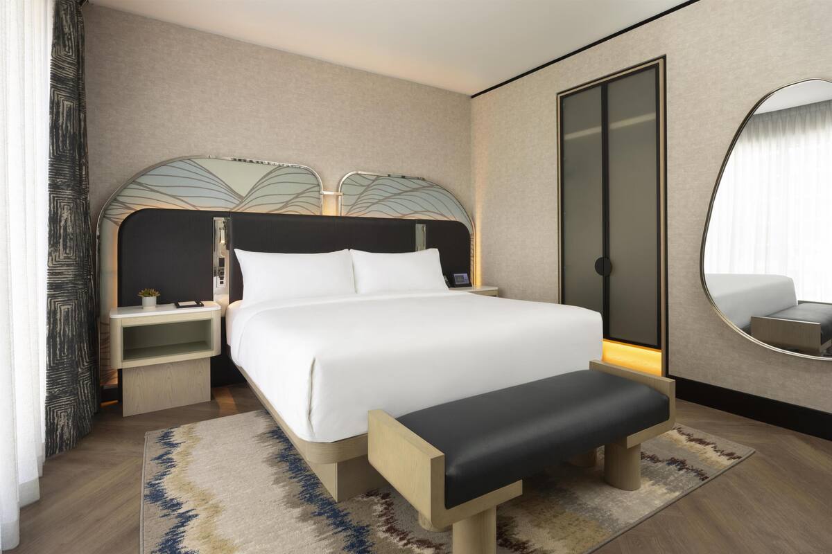 Una habitación de hotel moderna con una cama grande, mesitas de noche, un banco, un espejo y un armario.
