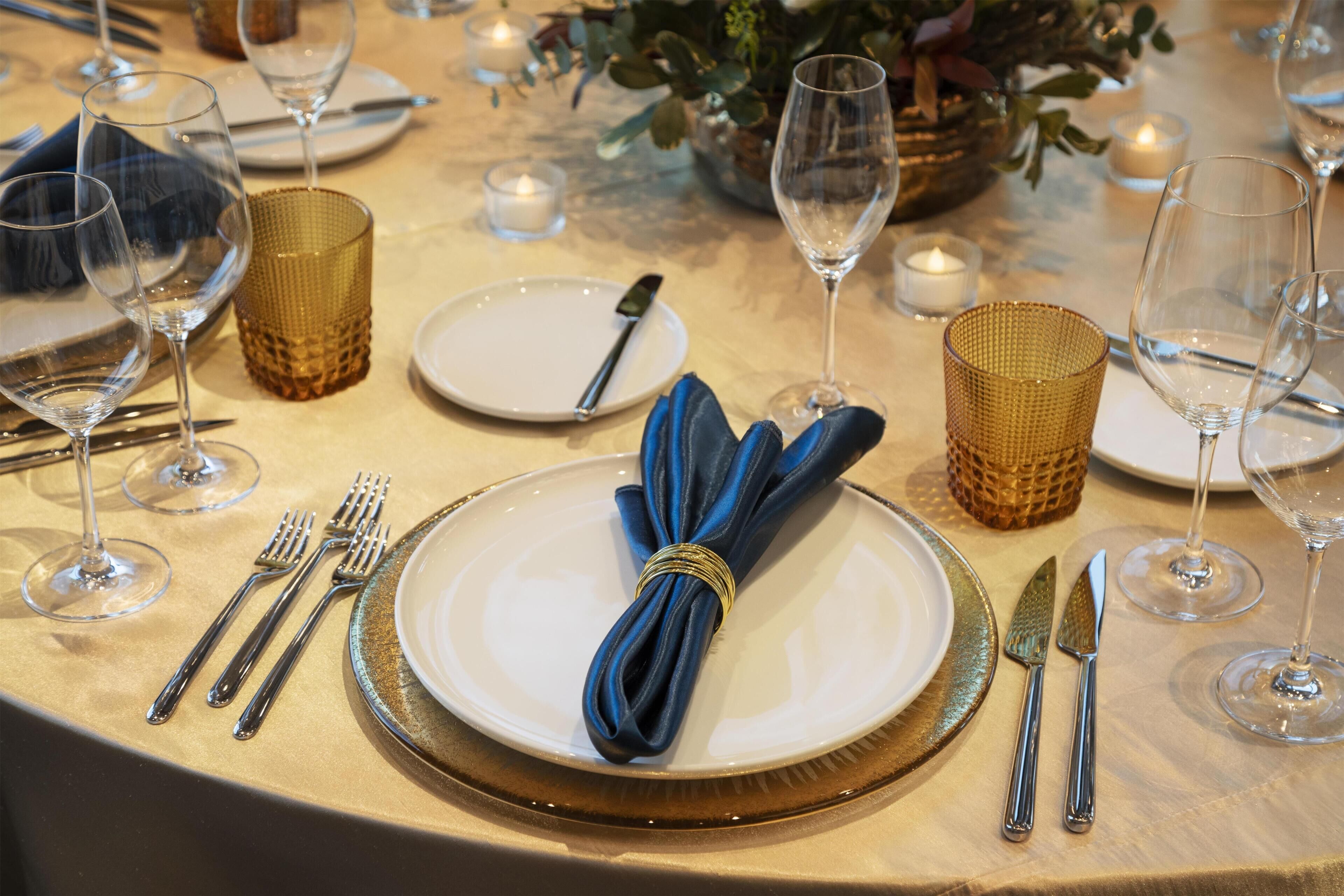 Una mesa dispuesta con platos, cubiertos y copas de vino, con un servilleta azul atada al plato.