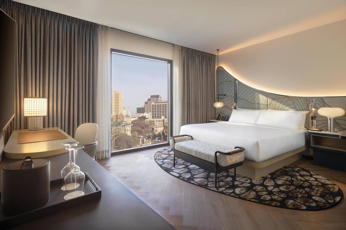 Una habitación de hotel moderna con una cama grande, un escritorio con lámpara, una silla y vistas a la ciudad.