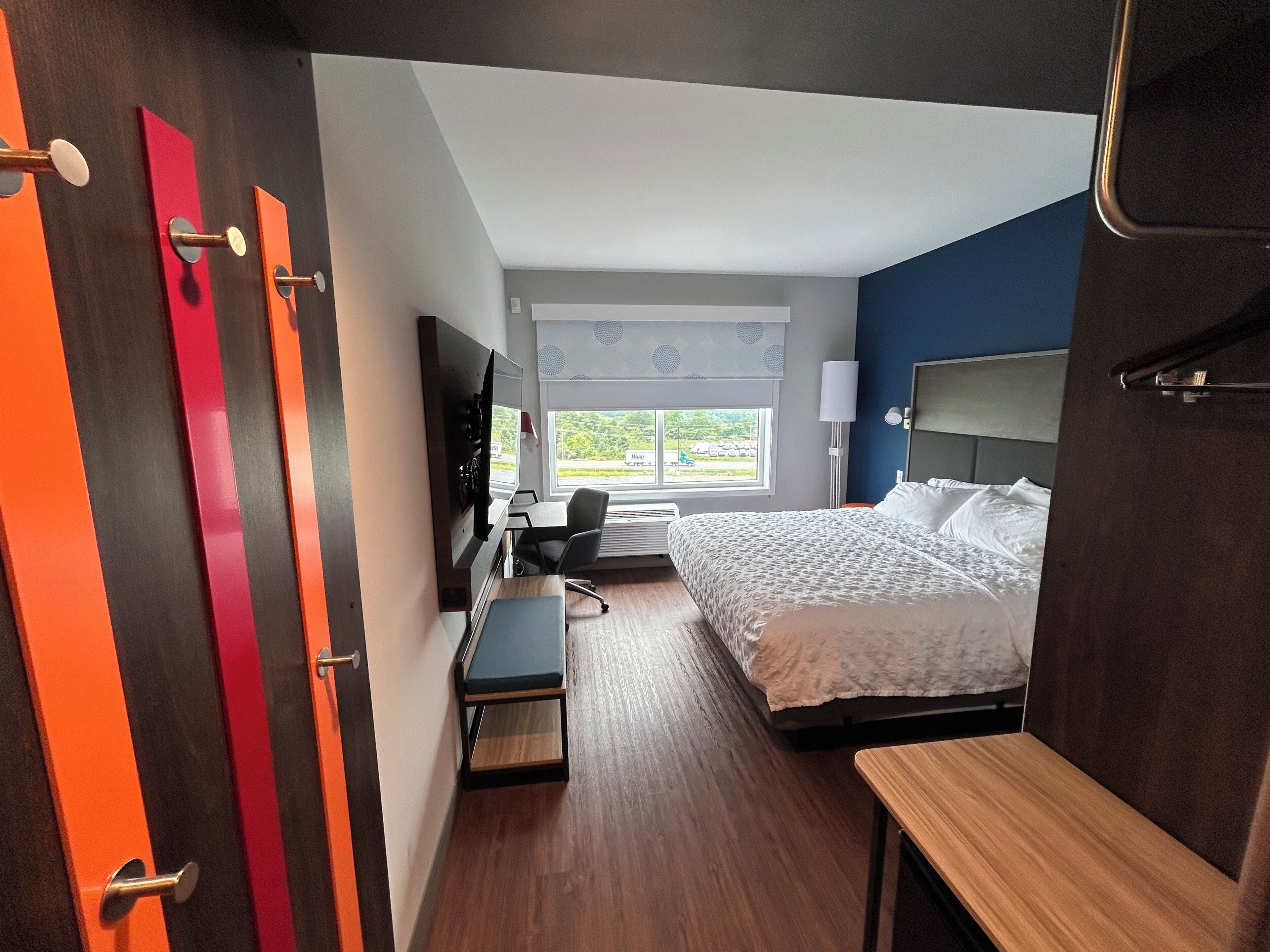 Une chambre d’hôtel comprenant un lit, un bureau avec une chaise, une télévision et une fenêtre donnant sur un espace verdoyant.