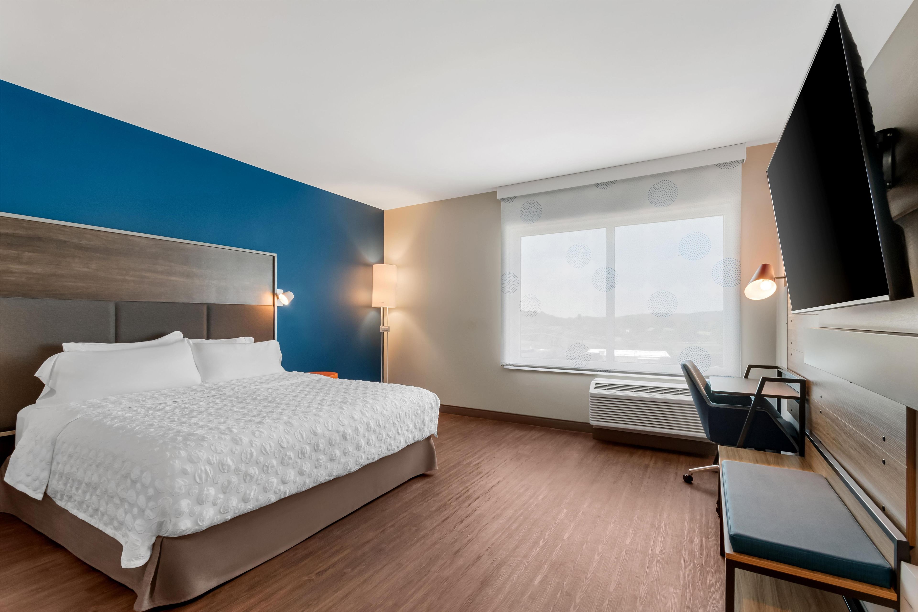 A hotel room with a bed, a desk, a chair, a TV, and a window with blinds.