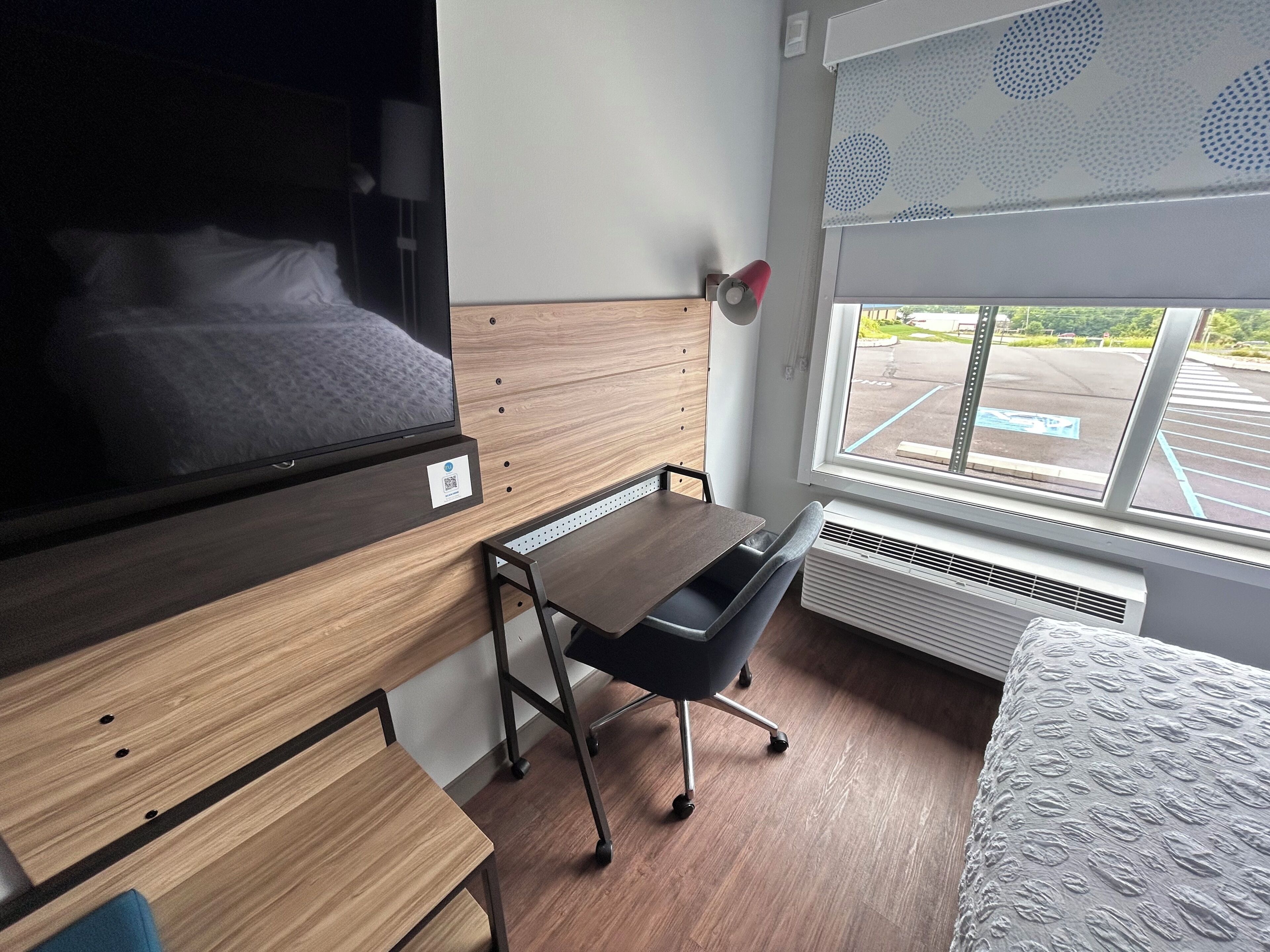 Ein Hotelzimmer mit Bett, Schreibtisch, Stuhl, Fenster mit Blick auf einen Parkplatz und einer Wandlampe.