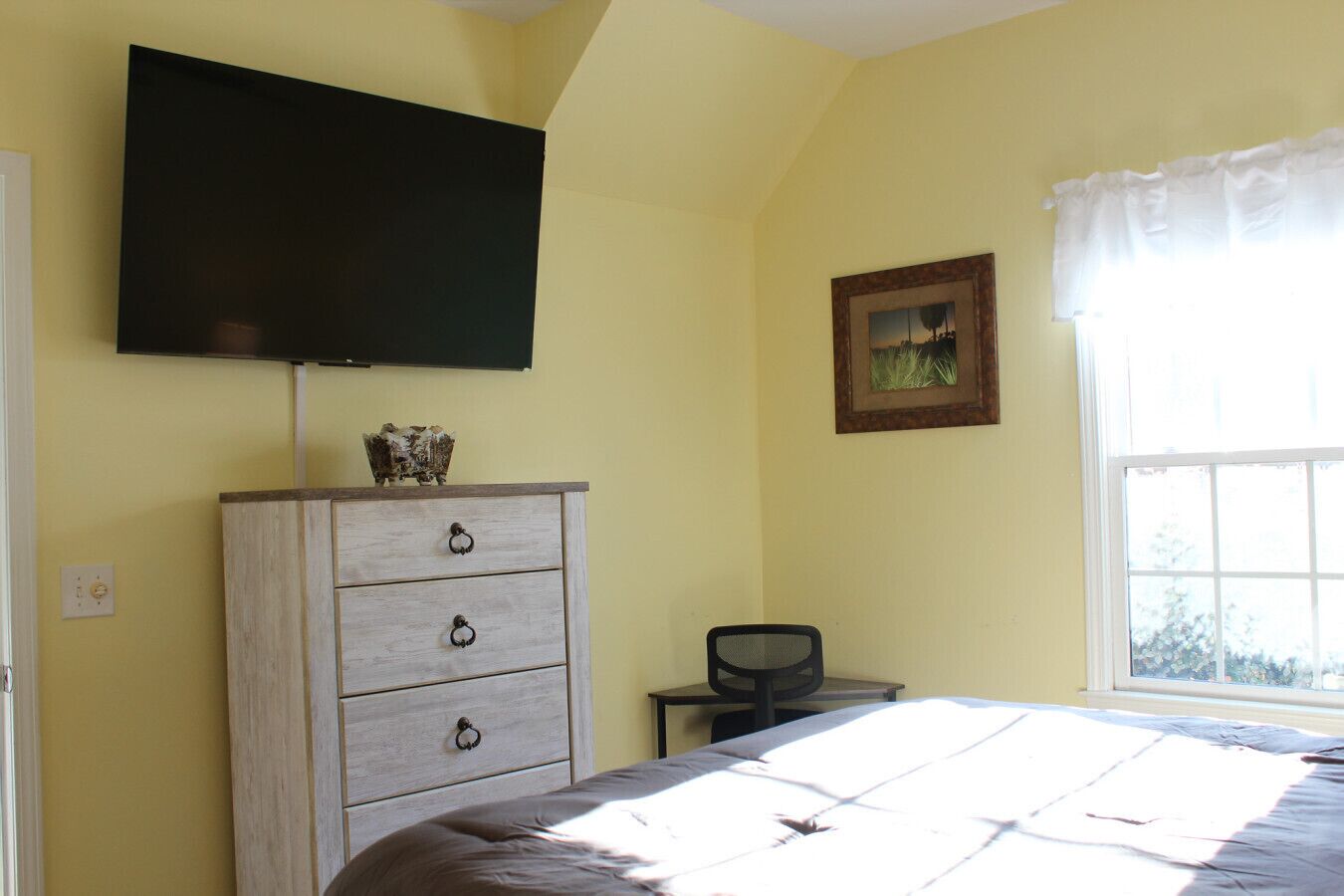 A bedroom with a bed, a dresser, a television mounted on the wall, and a window with curtains.