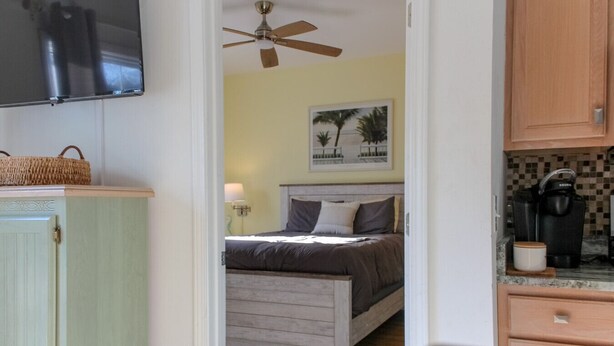 A bedroom with a bed, a ceiling fan, a nightstand, a lamp, and a wall-mounted TV. - The Villa at 205 Royal Poste Rd (Sunset Beach)