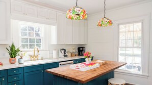 A kitchen with teal cabinets, a wooden island, wicker stools, a coffee maker, and a colorful stained glass pendant light. - Eclectic 1906 Farmhouse: 2 Acres, Fire Pit, Minutes to Lake Norman  & Charlotte (Huntersville)