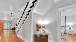 A spacious hallway with a wooden staircase, a wooden nightstand with decorative items, and a bed with a teal curtain in the adjacent room. - Eclectic 1906 Farmhouse: 2 Acres, Fire Pit, Minutes to Lake Norman  & Charlotte (Huntersville)