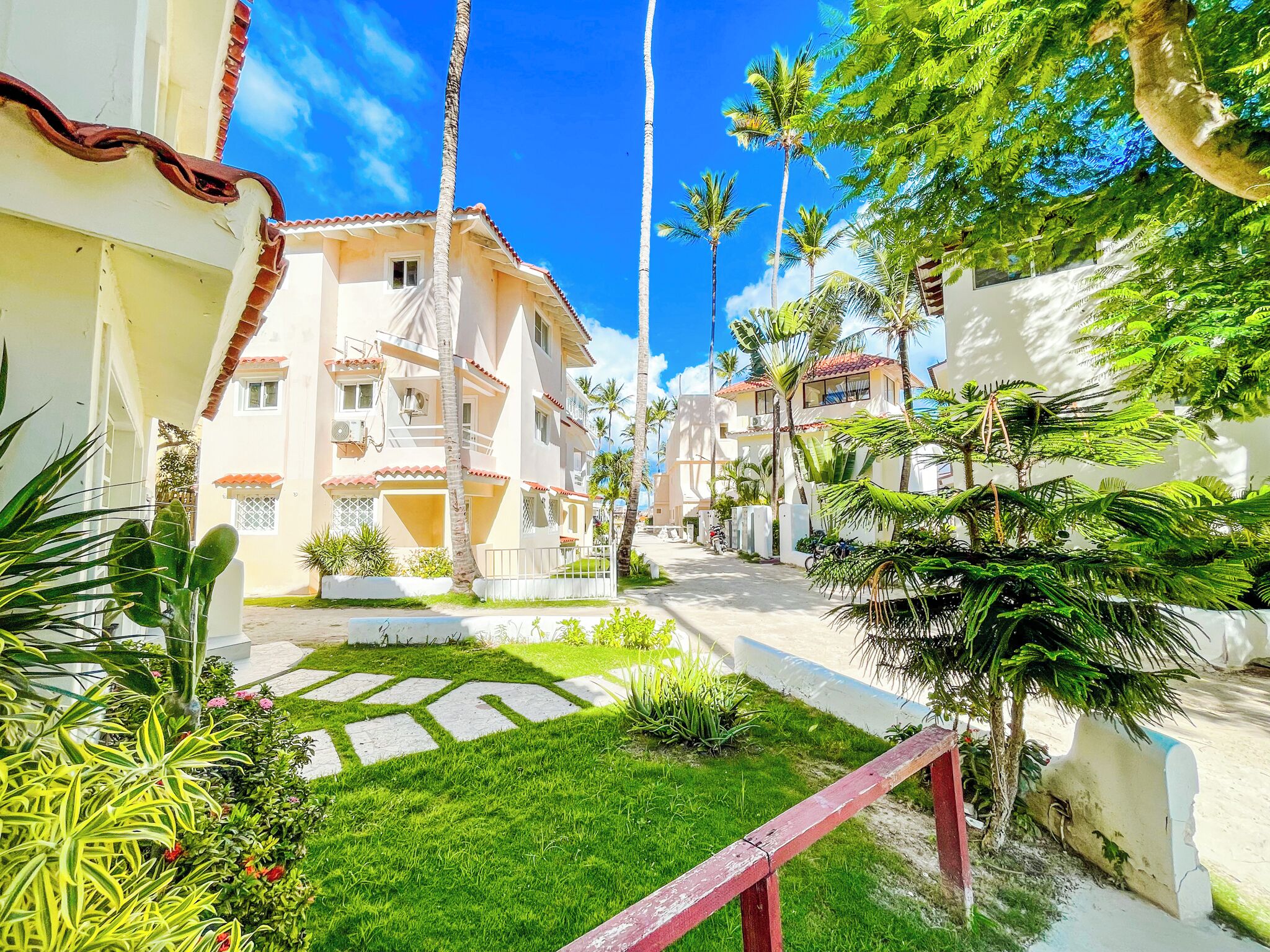 Zona residencial com calçada pavimentada, palmeiras e um edifício com telhado de telhas vermelhas.