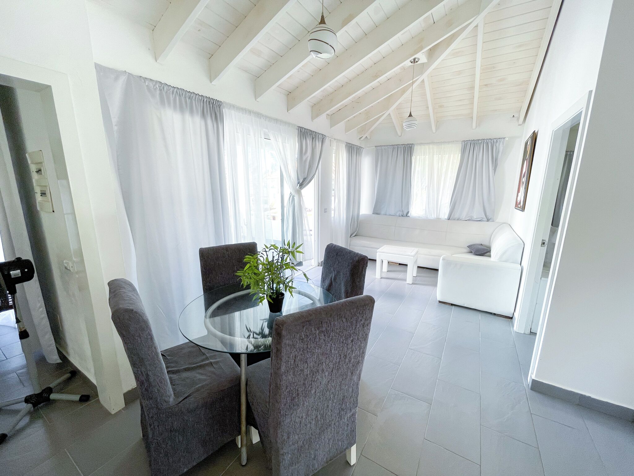 Uma sala de estar moderna com mesa de vidro, cadeiras cinzas, sofá branco e uma janela grande com cortinas.