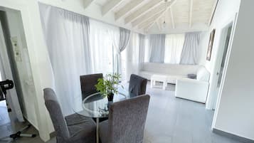 Uma sala de estar moderna com mesa de vidro, cadeiras cinzas, sofá branco e uma janela grande com cortinas.