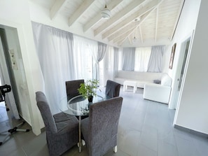 A modern living room with a glass table, grey chairs, a white sofa, and a large window with curtains. - BAVARO SEASIDE Villas BEACH HOTEL & SPA (Punta Cana)