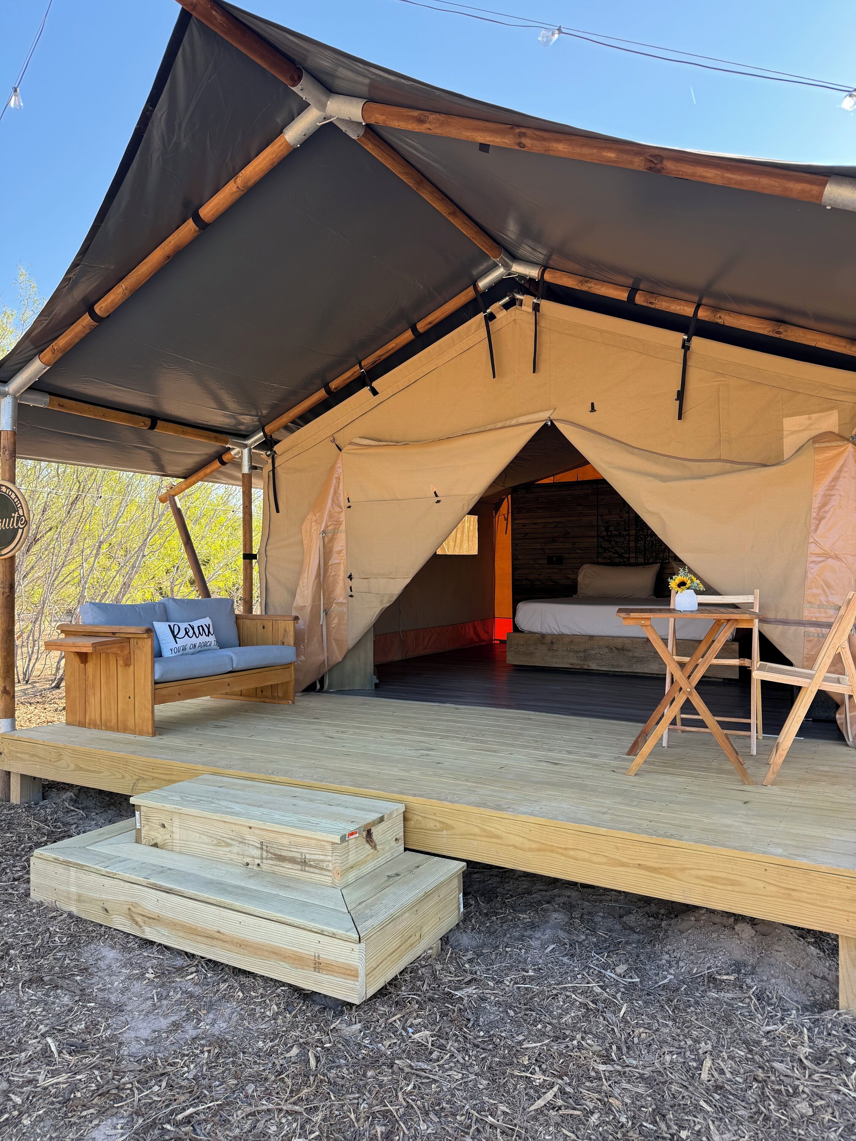 A tented accommodation with a wooden deck, a sofa with a 'Relax' pillow, and a table with a vase.