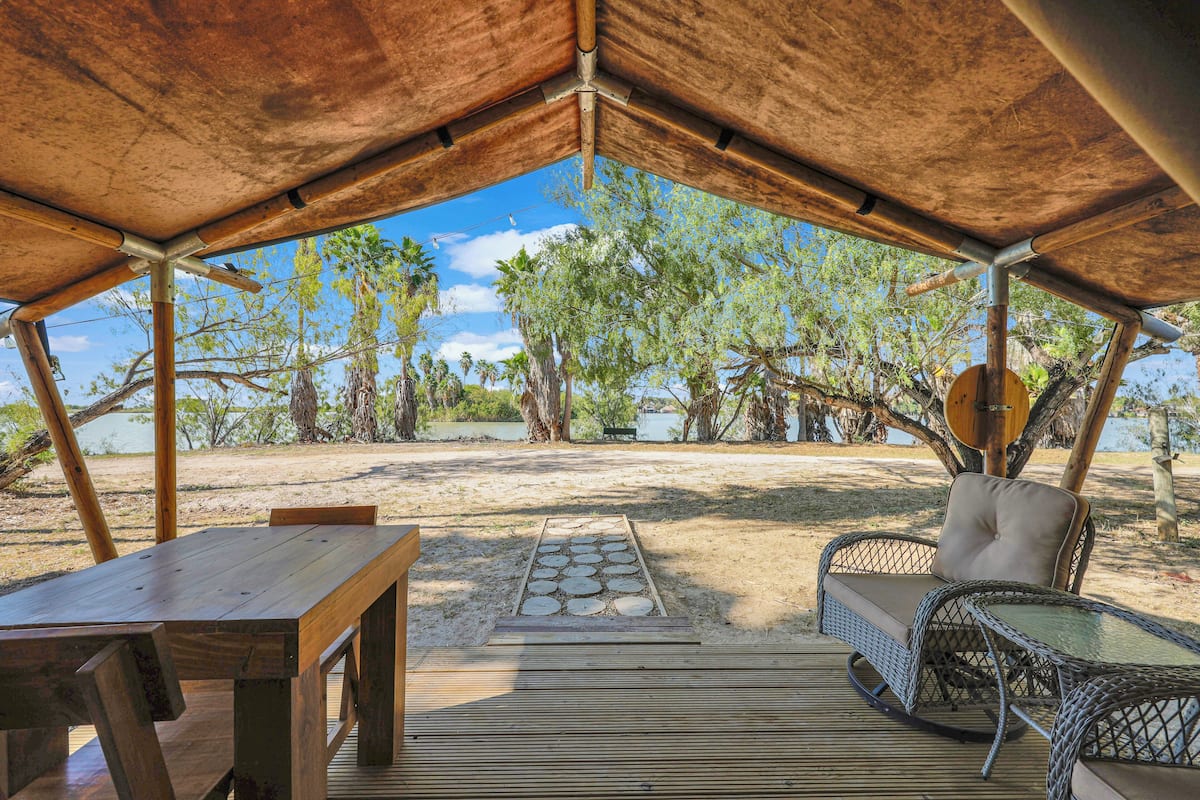 Un espacio al aire libre con mesa y sillas de madera, una silla tejida y vistas a árboles y agua.