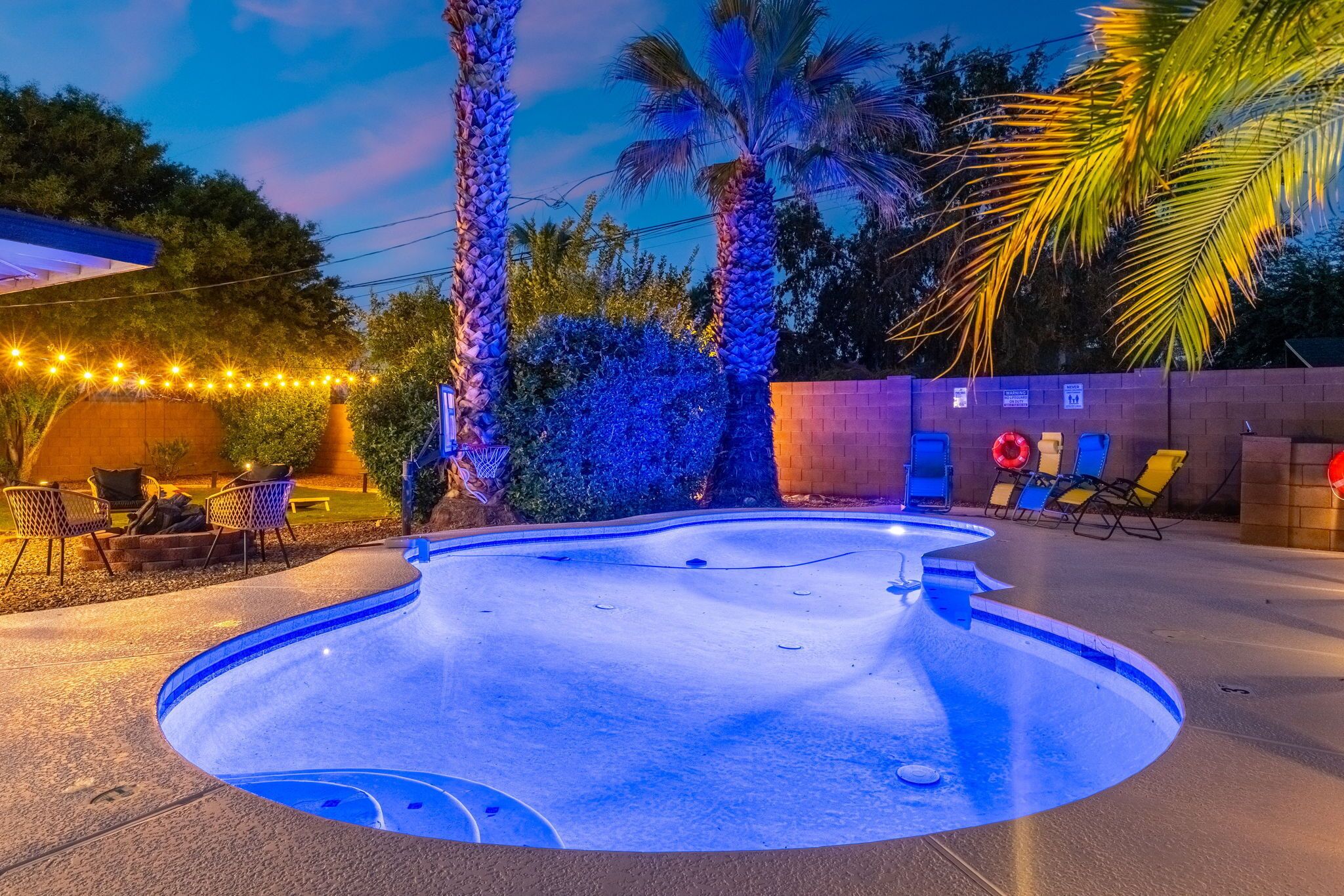 A swimming pool with a curved edge, surrounded by palm trees and outdoor furniture.