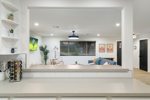 A modern living room with a sofa, a coffee table, and a dining area with a counter. There are shelves with plants and a hanging light fixture.