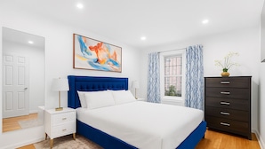 A bedroom with a large bed, a nightstand, a dresser, a window with curtains, and a painting on the wall.
