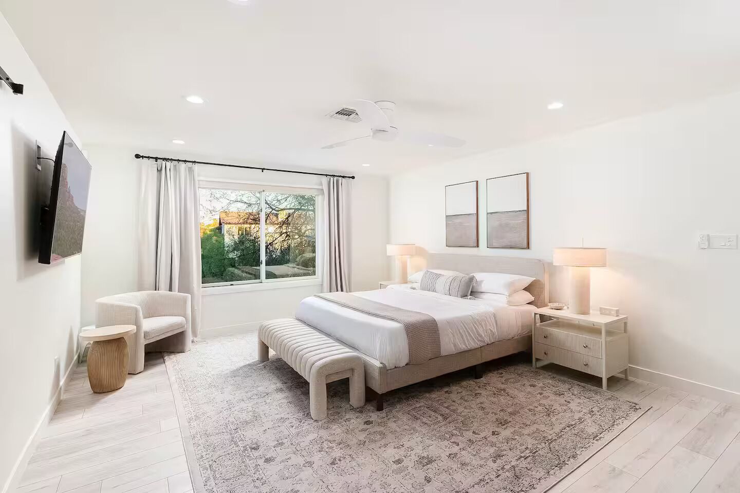 A modern bedroom with a large bed, bedside tables, a chair, a TV, and a window with curtains.