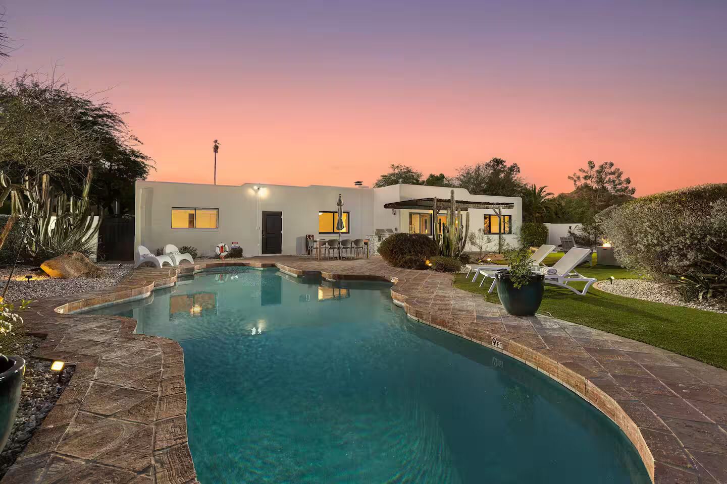 A swimming pool with a stone border, a patio area with chairs, and a building with windows and a door.