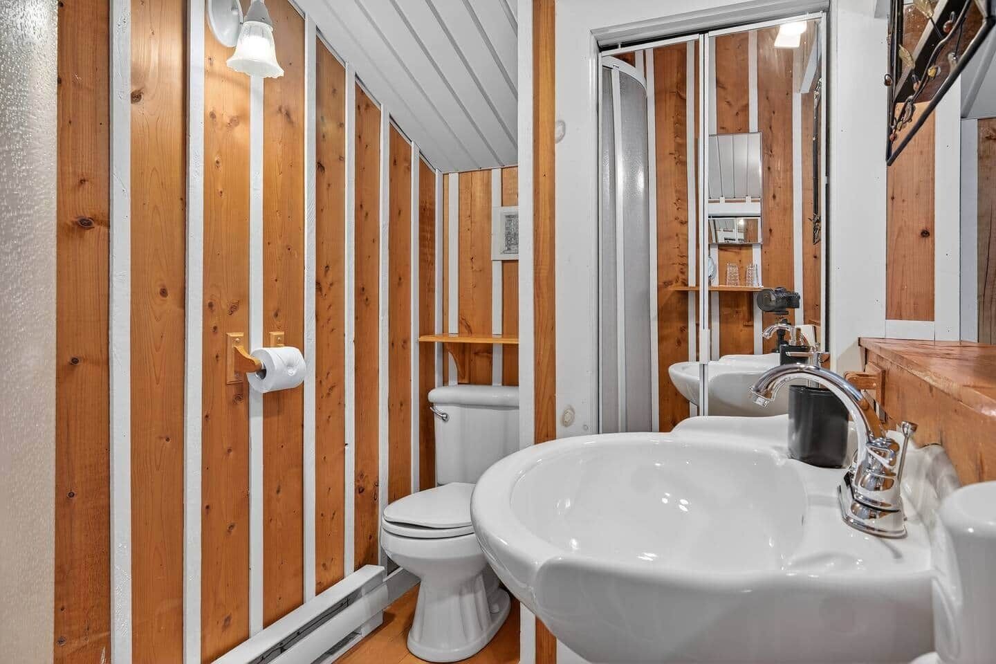Un baño con paneles de madera, un inodoro blanco, un lavabo con espejo y una zona de ducha.