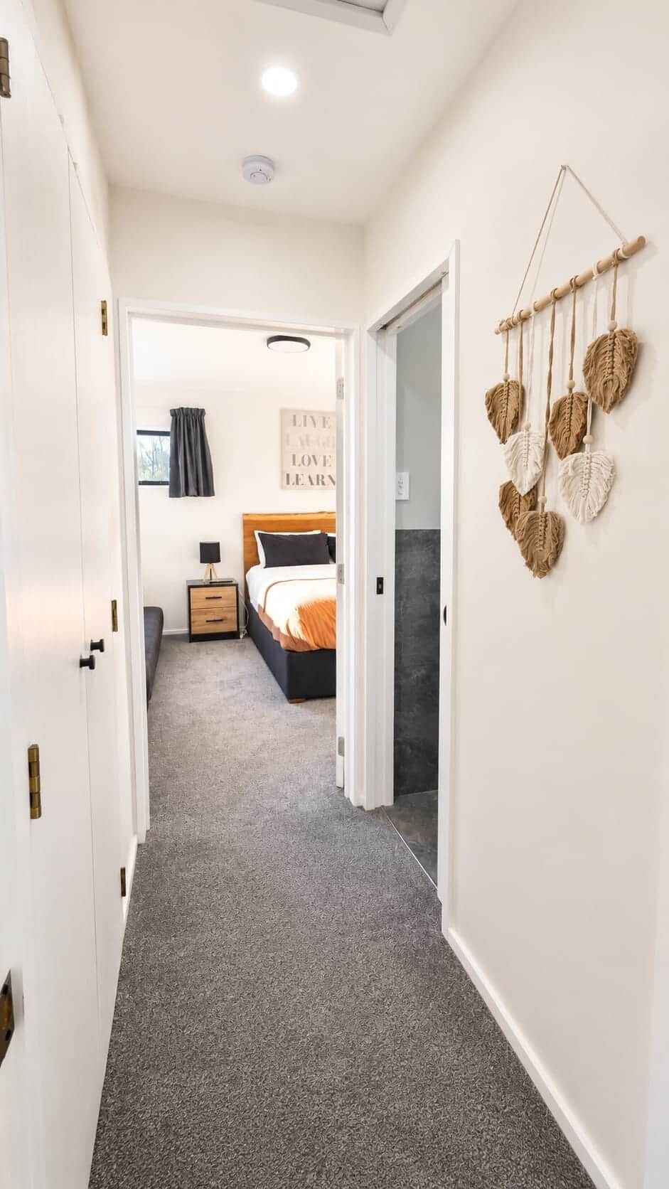Un couloir menant à une chambre à coucher, où se trouvent un lit, une table de chevet et une décoration murale en macramé.