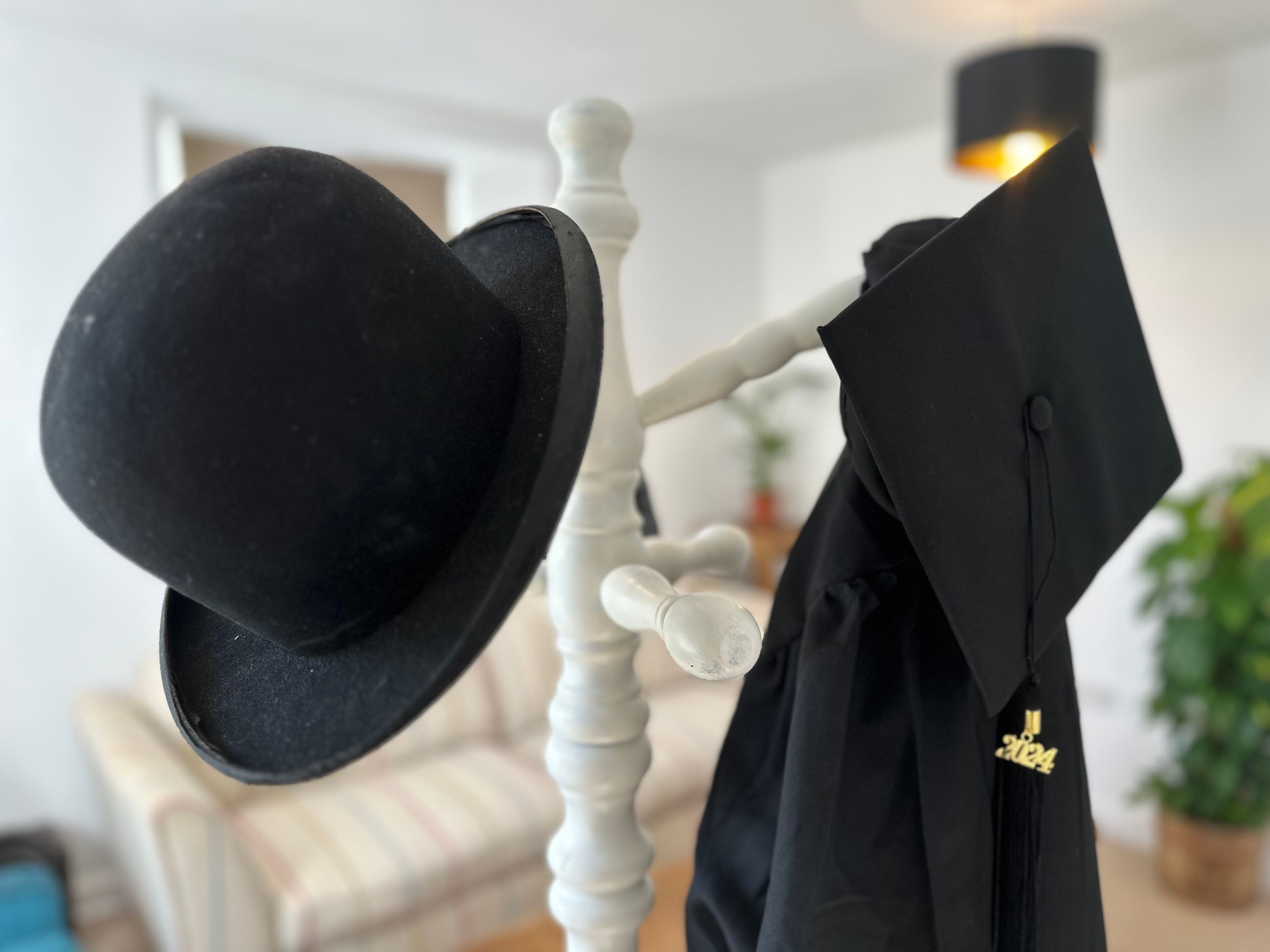 Une toque et une toge de remise de diplômes noires sont suspendues à un présentoir blanc dans une pièce où l’on aperçoit une chaise blanche et une lampe à l’arrière-plan.