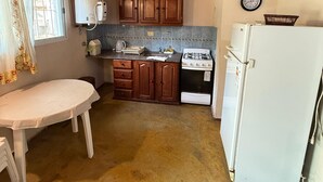 A compact kitchen with wooden ceiling beams, a white table, wooden cabinets, a gas stove, and a white refrigerator. - Cabañas Sol Mediterraneo (Cosquín)