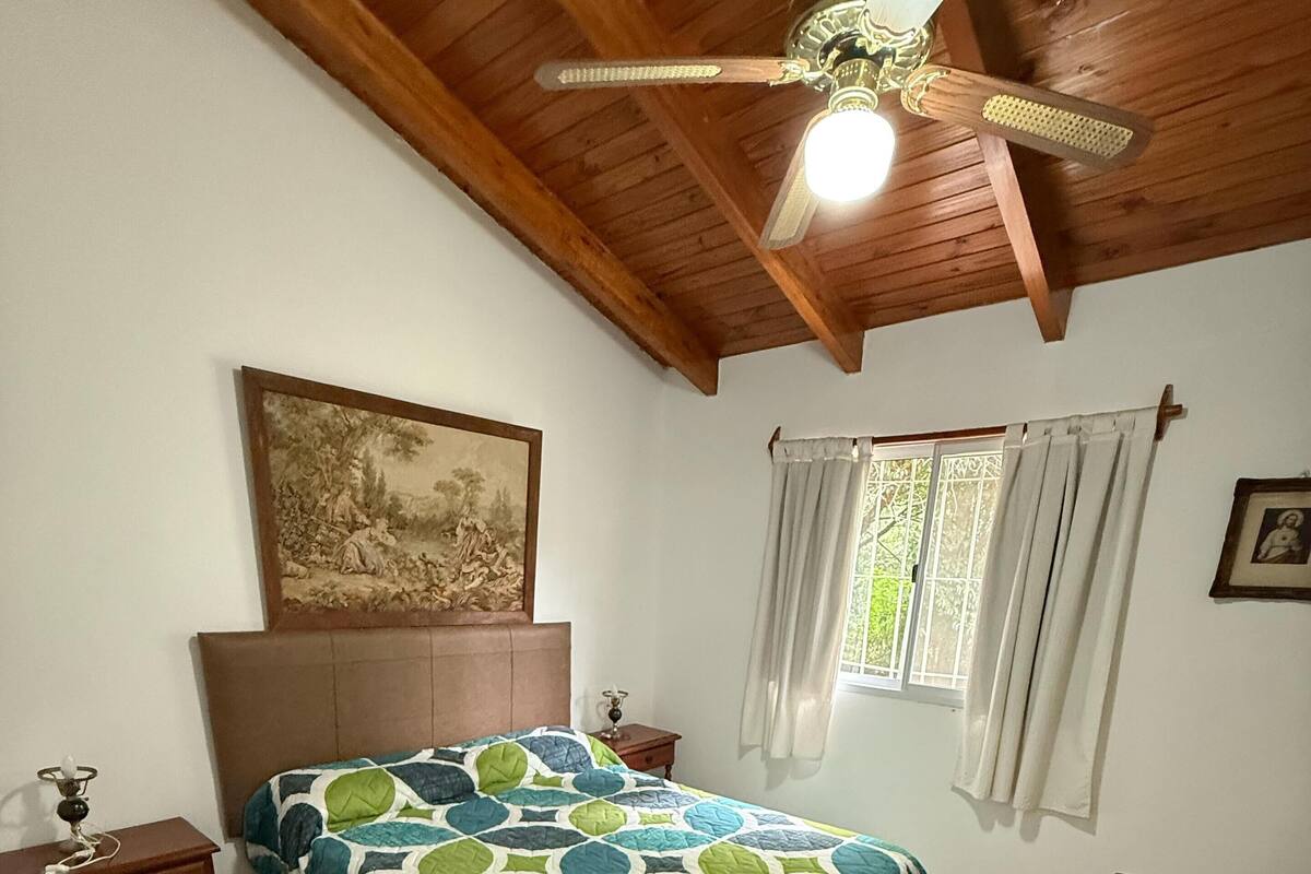 A bedroom with a bed, a wooden headboard, a ceiling fan, a window with curtains, a nightstand, and a dresser.