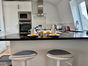 Una cocina moderna con una isla central, dos taburetes de bar y alimentos para el desayuno, como cruasanes y jugo de naranja.