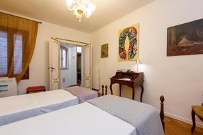 Quarto de hotel com duas camas, uma escrivaninha, uma cadeira, um radiador e um quadro na parede.