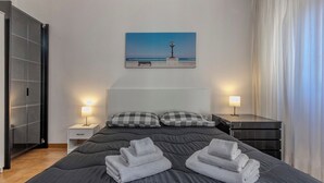 A hotel room with a bed, two bedside tables, a nightstand, a painting of a beach scene, and a large window with curtains. - Andrea's home Bari (Bari)