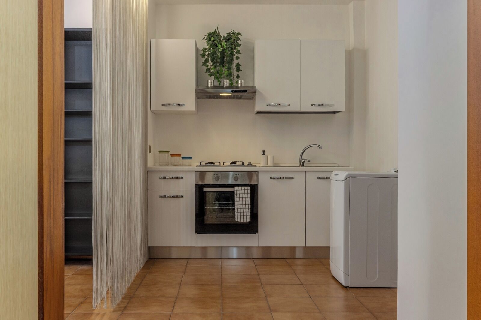 Une cuisine compacte avec des meubles blancs, un four encastré, un évier et une petite plante posée sur le plan de travail.