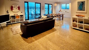 Un salon moderne avec un grand canapé noir, un téléviseur à écran plat et un coin repas comprenant une table et des chaises. De grandes fenêtres offrent une vue sur un port de plaisance.