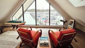 Une pièce dotée d’une grande baie vitrée donnant sur un port de plaisance, de deux fauteuils rouges, d’une petite table et d’un tableau encadré accroché au mur.