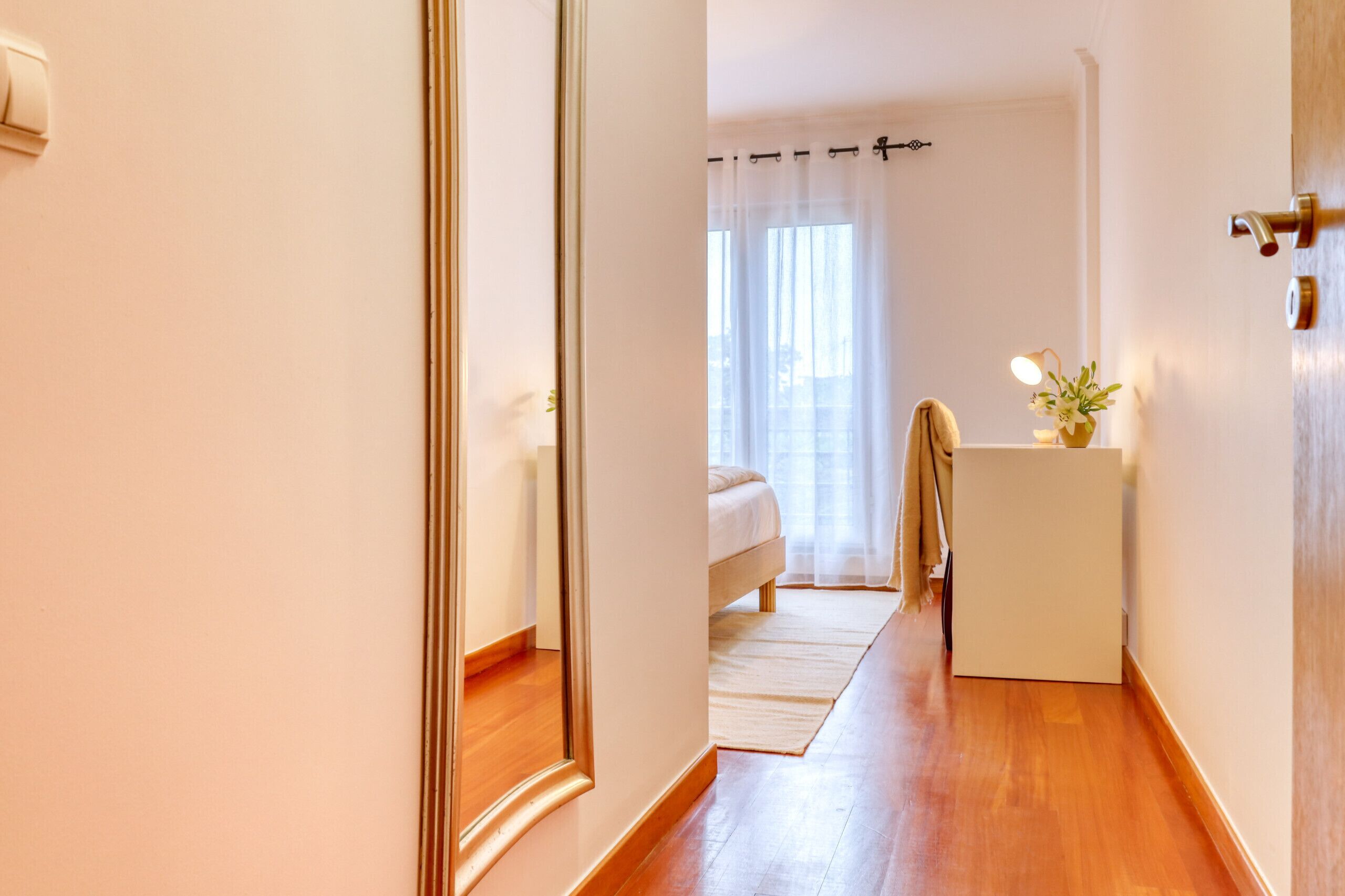 Un couloir mène à une chambre à coucher qui contient un lit, une petite table avec une plante et un miroir sur le mur de gauche.