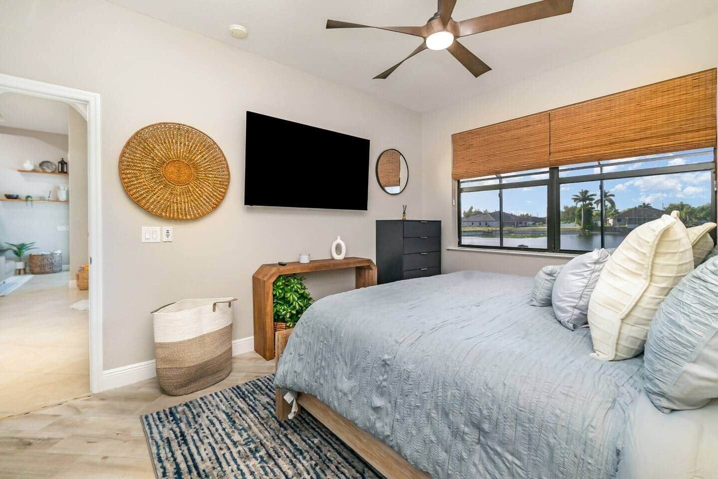 A bedroom with a bed, a TV, a window with a view, a ceiling fan, and a wall-mounted shelf.