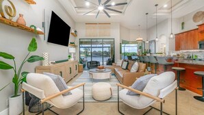 A spacious living area with a large TV, comfortable seating, a kitchen with bar stools, and a view of the outdoors. - Sho Me Sunshine - Lake Front w/ Southern Exposure (Cape Coral)