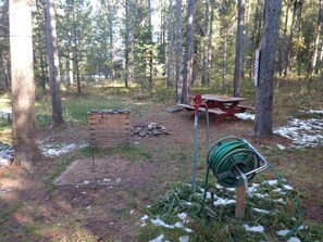 A forested campsite with a picnic table, a wooden bench, and a green garden hose.